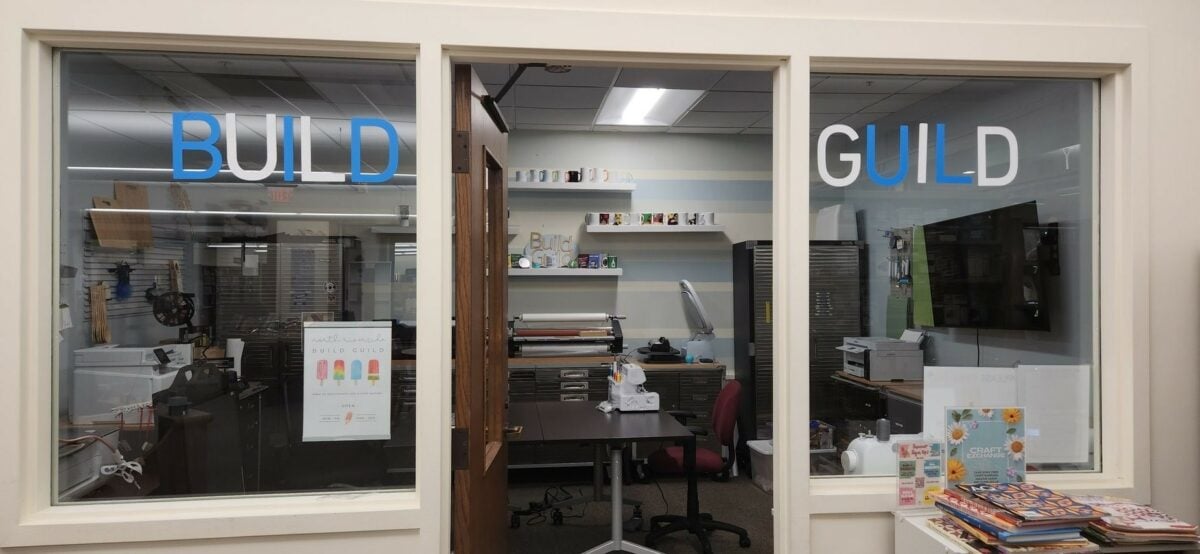 Photo of the Build Guild makerspace with an open door and the worlds "BUILD GUILD" in blue and white vinyl on the windows on either side of the door. Inside is a sewing machine, large laminator, 3D printer, wall mounted TV monitor, printer and cabinets.