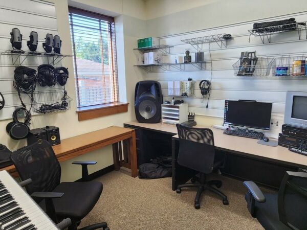 Interior photo of the Studio 2400 room showing a keyboard, electronic drums, ukulele, guitar, microphones and more for the music recording and a computer, vhs to dvd converter and additional converting equipment for the digitization side of the room. There are 3 chairs, a desk and a table.